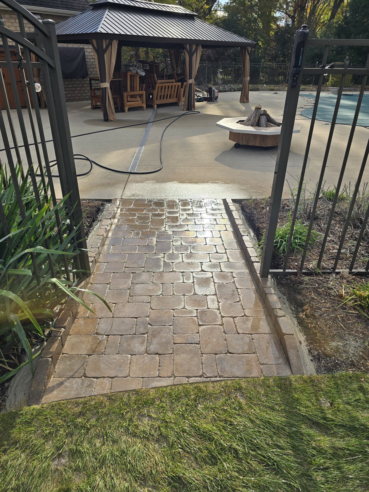 A paver walkway through an open garden gate leading to a backyard patio and pavilion