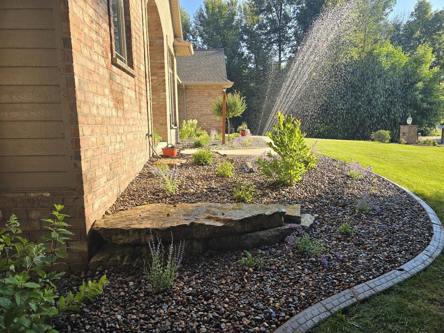Fresh plantings and a broad accent boulder in a curved river rock bed beside a brick home